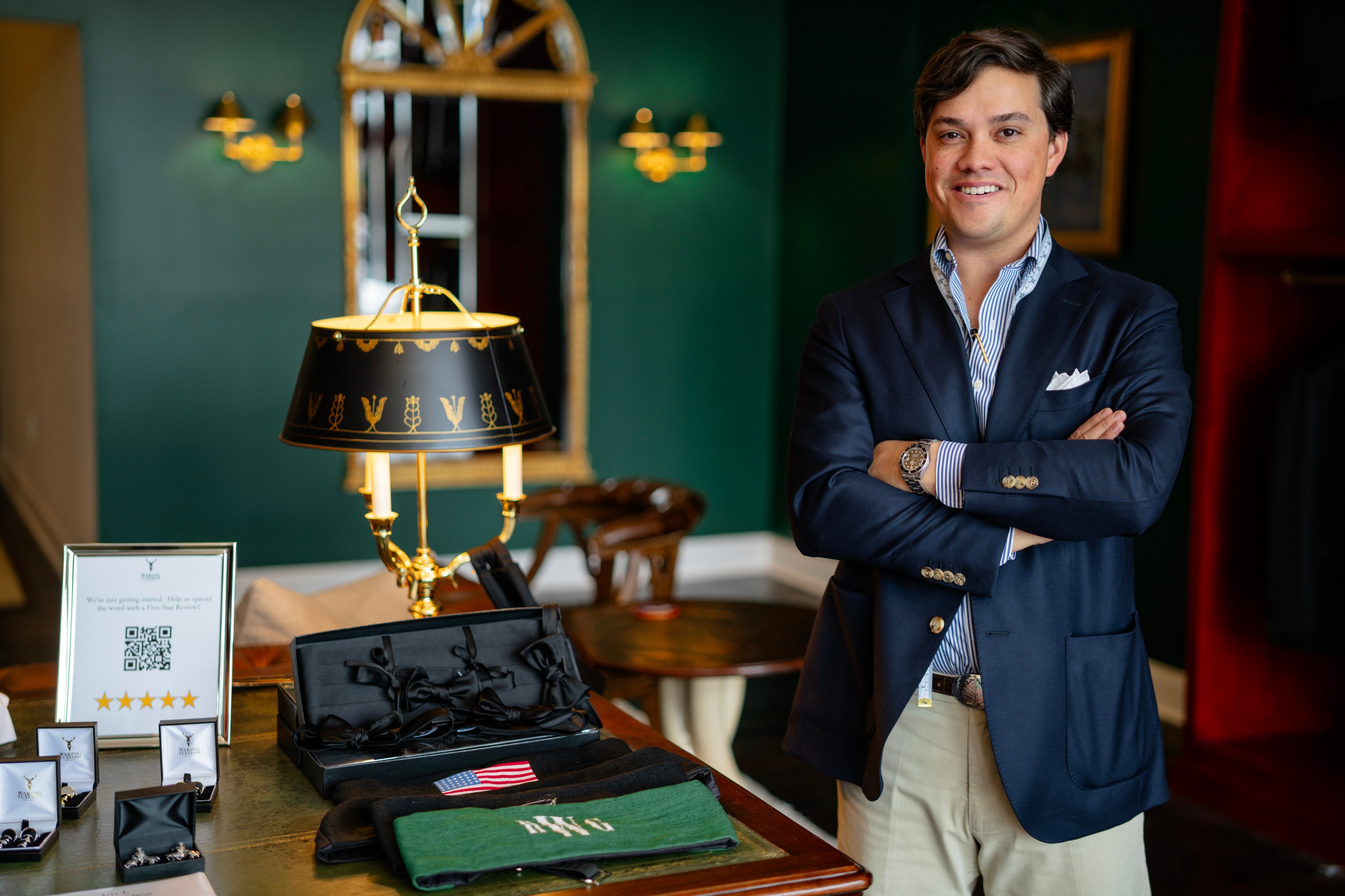 Houstoun Waring inside Waring Tuxedos, a Fort Worth tuxedo shop, standing beside formalwear accessories in a green-walled, mahogany-shelved showroom.
